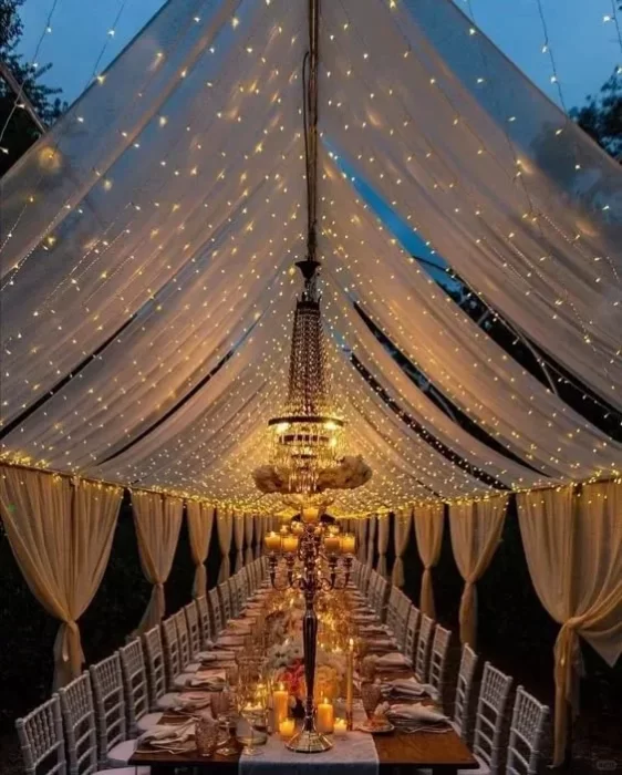 string light canopy over a long wedding banquet table, with draped fabric and a chandelier, creating a warm, romantic outdoor evening reception setting.