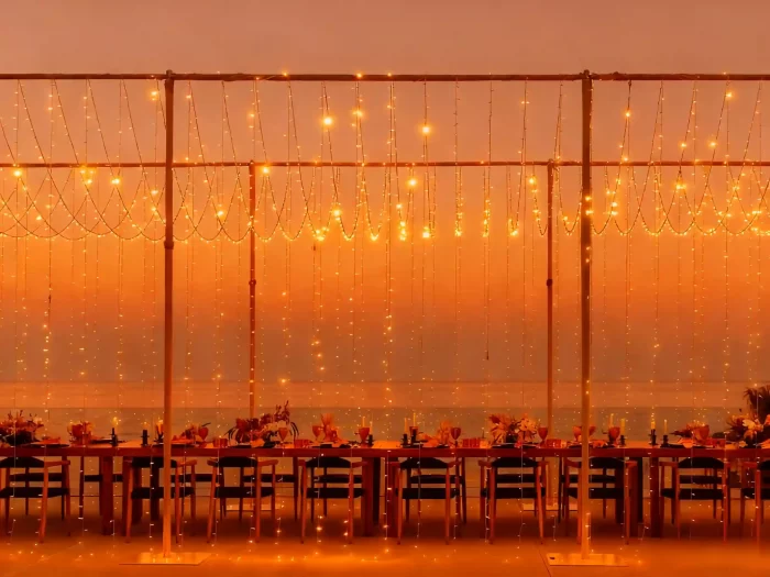string light curtain backdrop framing a long outdoor banquet table by the sea at sunset; warm amber LEDs in scalloped rows create romantic event lighting.