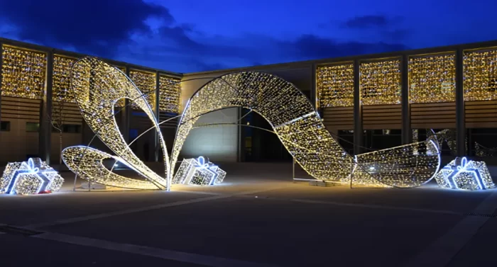 A large 3D LED motif light sculpture shaped like a ribbon with glowing gift boxes, illuminated at night. Perfect for holiday displays in public spaces.