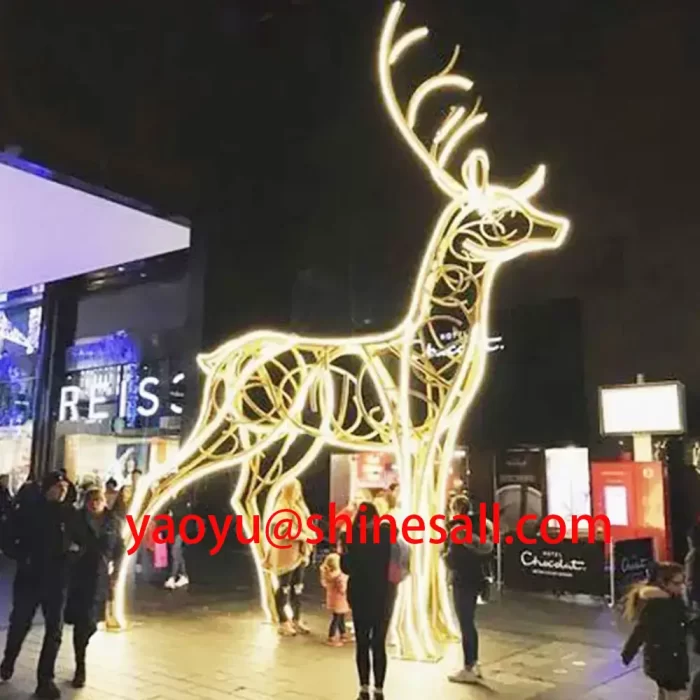 A giant 3D reindeer light sculpture, illuminated with warm golden LEDs, stands as a centerpiece in a busy city shopping plaza at night. The sculpture features an intricate scrollwork body, and families with children are seen admiring this festive outdoor Christmas decoration in front of a REISS store.