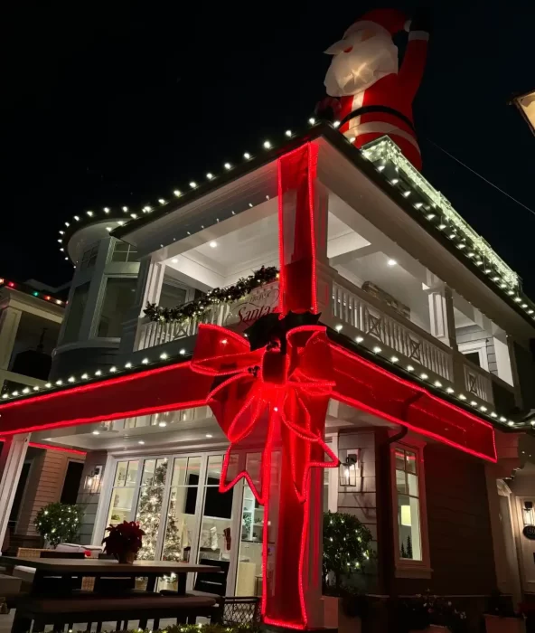 Two story house decorated for Christmas with a giant red LED ribbon and bow across the facade and an inflatable Santa on the roof