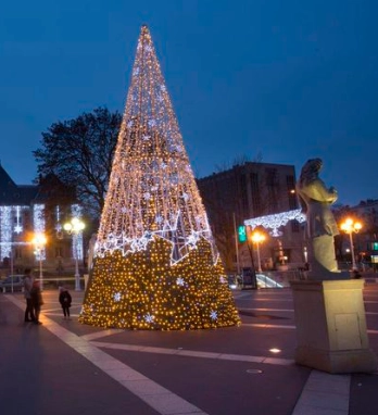Cone-shaped LED Christmas tree installation in a public plaza at dusk, wrapped in warm white lights.
