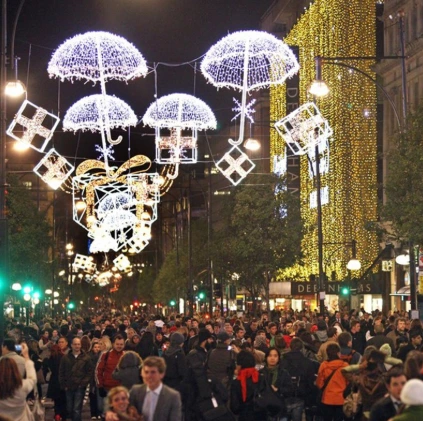 Holiday street canopy with glowing umbrella and gift-box light motifs above a crowded city shopping avenue at night.